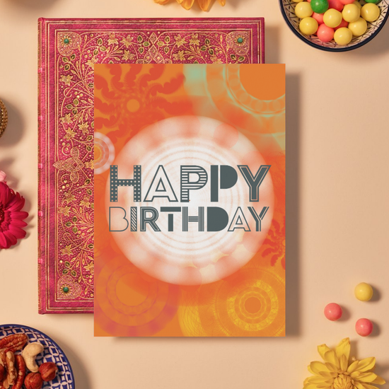 Card with bold dark grey text reading ‘HAPPY BIRTHDAY’ in a mix of dotted, striped, and outlined geometric fonts. The background features vibrant orange and yellow mandala patterns made of circular and spiral geometric shapes radiating outward, with a glowing white mandala at the center, creating a festive and energetic design.