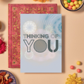 Card with bold dark grey text reading ‘THINKING OF YOU’ in a mix of outlined and striped geometric fonts. The background features soft blue, cream, and peach mandala patterns made of layered circular and floral geometric shapes, creating a calm, reflective, and comforting design.