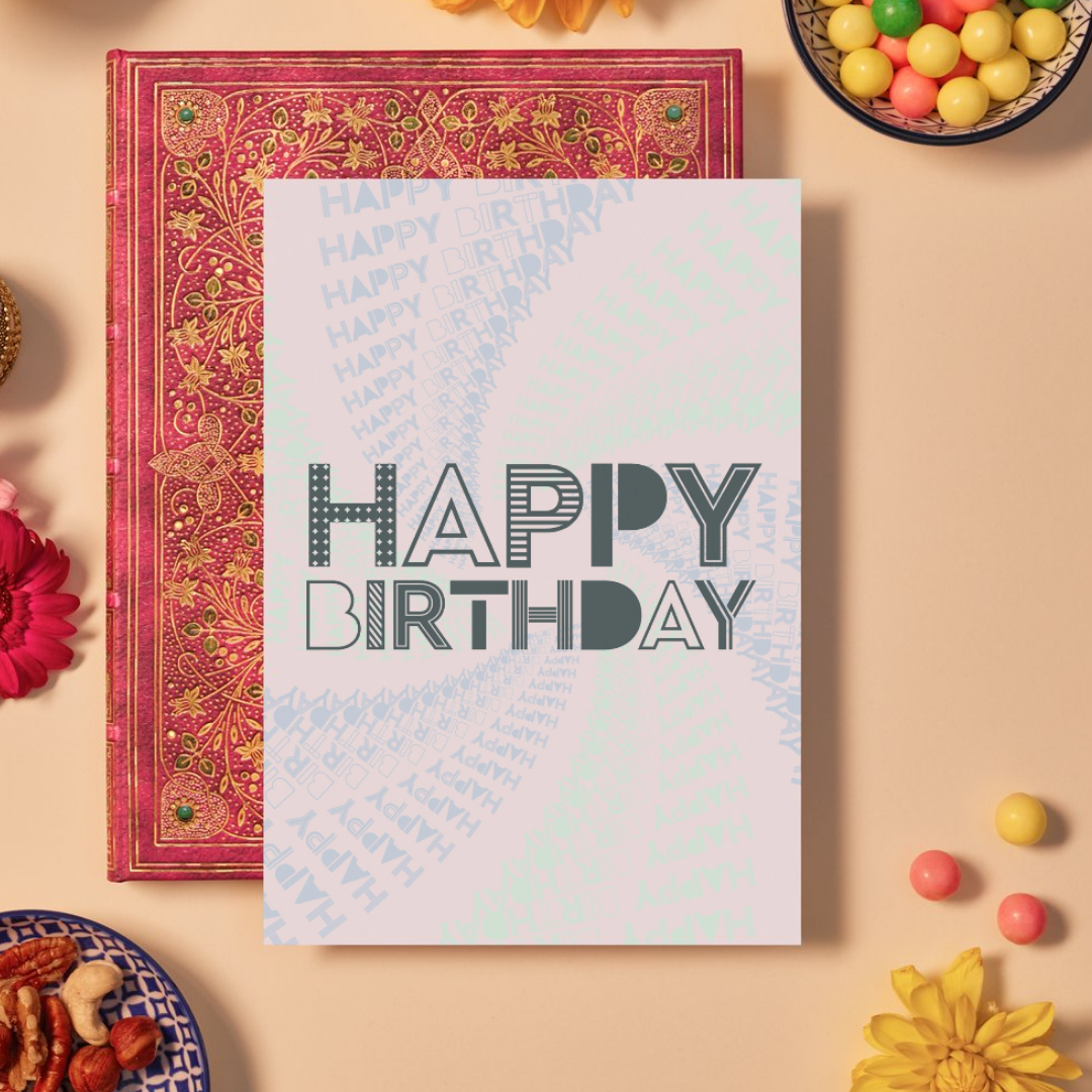 Card with bold dark grey text reading ‘HAPPY BIRTHDAY’ in a mix of striped, dotted, and outlined geometric fonts. The background features a soft pink and mint green mandala pattern formed by repeating the phrase ‘HAPPY BIRTHDAY’ in varying sizes and shades, spiraling outward to create a festive, energetic design.