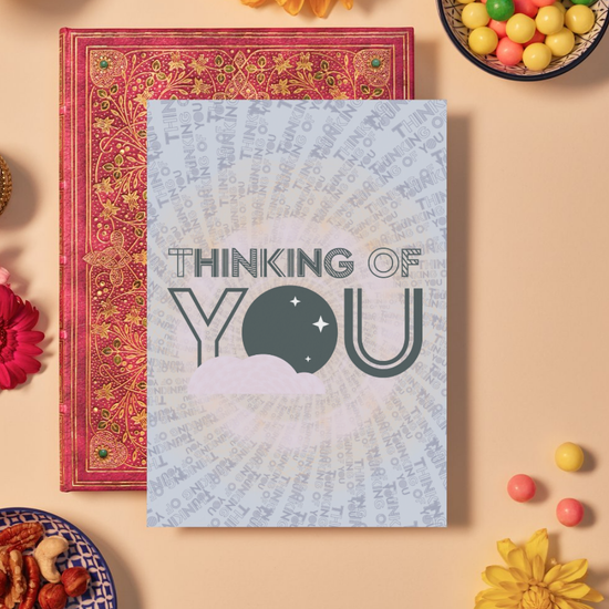 Card with bold dark grey text reading ‘THINKING OF YOU’ in a mix of outlined and striped geometric fonts. The background features a mandala-like pattern made from radiating repetitions of the phrase ‘THINKING OF YOU’ in various sizes and shades of grey and peach. In the center, a dark grey circle with white star shapes and a soft pink cloud adds a calm, reflective touch.