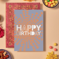 Birthday card with bold white text reading ‘HAPPY BIRTHDAY’ in a mix of playful geometric fonts, some striped, dotted, or outlined. The background features a textured orange mandala pattern radiating outward on a soft lavender base, creating a lively and celebratory look.