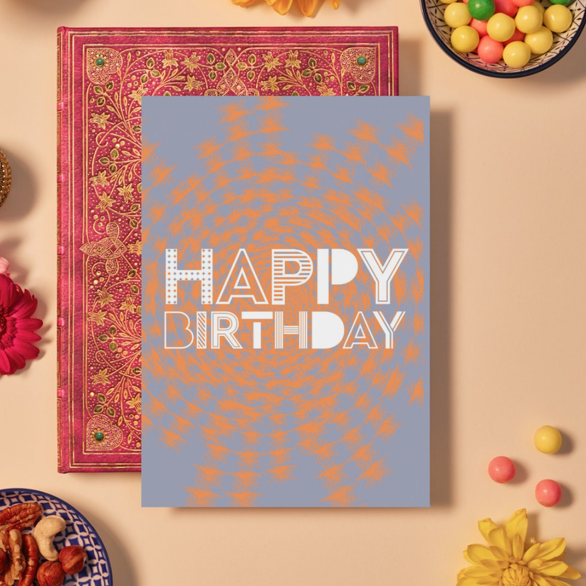 Birthday card with bold white text reading ‘HAPPY BIRTHDAY’ in a mix of playful geometric fonts, some striped, dotted, or outlined. The background features a textured orange mandala pattern radiating outward on a soft lavender base, creating a lively and celebratory look.
